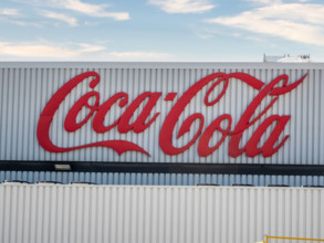 Ein großes, rotes Coca-Cola-Logo prangt auf einem weißen Gebäude mit vertikalen Streifen unter einem teilweise bewölkten Himmel.