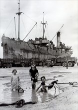 French children playing in a pool on the beach at Arromanches while a ship discharges its cargo to