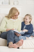 Grandmother reading to young granddaughter on sofa.