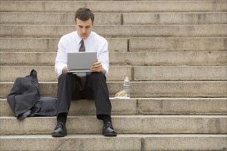 Young adult male sitting outdoors on stairs with laptop.