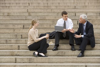 Three business people talking outdoors on stairs with laptop.