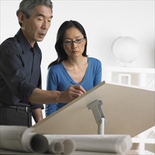 Asian businessman and businesswoman at drafting table with blueprints.
