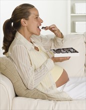 Pregnant woman on sofa eating box of chocolates.