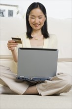 Asian woman with credit card sitting on floor using laptop .