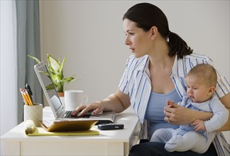 Mother typing on laptop and holding baby.