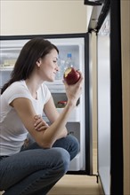 Woman eating apple in front of open refrigerator.