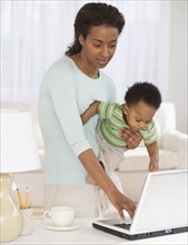 Mother holding baby and typing on laptop.
