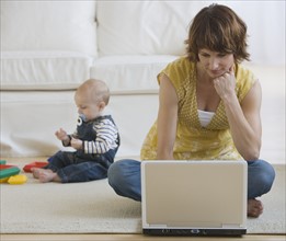 Mother using laptop while baby plays.