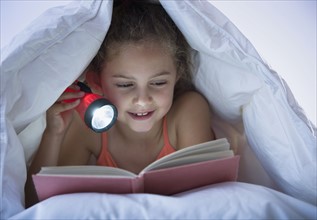 Girl reading with flashlight under blanket.