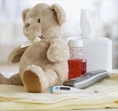 Teddy bear, medication, thermometer and telephone on blanket.