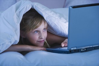 Teenaged girl under blanket looking at laptop.