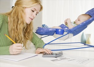 Mother using calculator with baby in seat on table. Date : 2006