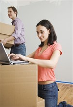 Woman using laptop while man carries box in new house. Date : 2006