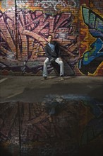 Asian man sitting in front of graffitied wall. Date : 2007