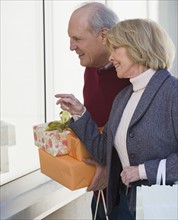 Senior couple holding gifts next to window.