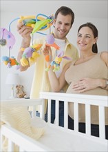 Pregnant Hispanic couple holding mobile over crib.