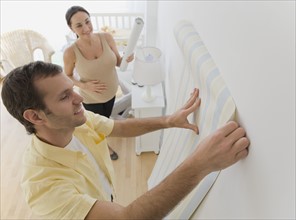 Pregnant Hispanic couple hanging wall paper in nursery.