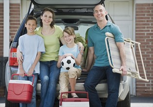 Family holding coolers and beach chair behind car.