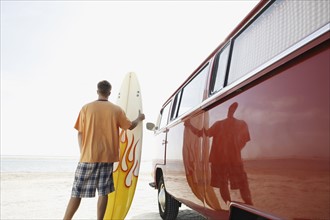 Man holding surfboard next to van on beach. Date : 2008