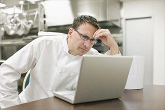 Frustrated chef looking at laptop in kitchen. Date : 2008