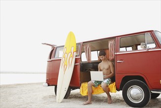 Young surfer using laptop in van on beach. Date : 2008