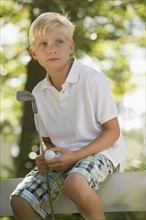 Boy sitting on fence with golf club and ball. Date: 2008