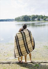 Couple wrapped in blanket looking out at lake. Date: 2008