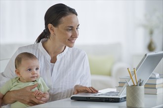 Mother working on laptop and holding baby son.