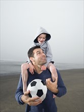 Father carrying son on shoulders and holding soccer ball on beach. Date : 2008