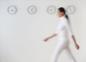 Business woman walking along wall with clocks, blurred motion.