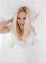 Portrait of young woman messing about under pristine white pillows. Photographe : Jamie Grill