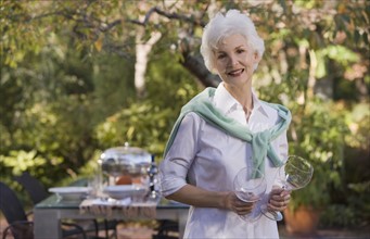 Senior woman standing in garden holding wineglasses. Photographe : mark edward atkinson