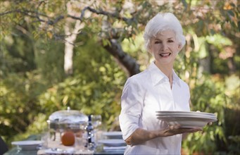 Senior woman standing in garden holding stack of plates. Photographe : mark edward atkinson