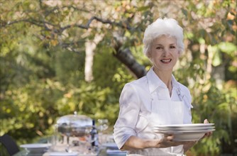 Senior woman standing in garden holding stack of plates. Photographe : mark edward atkinson