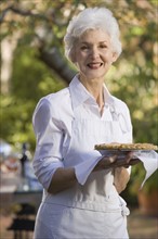 Senior woman standing in garden holding pie. Photographe : mark edward atkinson