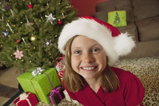 Girl (9) wearing Santa hat sitting in front of Christmas tree, portrait. Photographe : Sarah M.