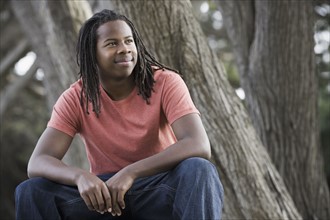 Teenage boy (16-17) with dreadlocks, sitting in park. Photographe : PT Images