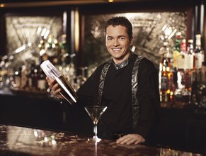 Young man standing behind bar shaking cocktail shaker, portrait. Photographe : Stewart Cohen