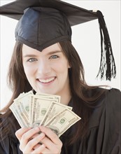 Portrait of young woman in graduation gown holding dollars. Photographe : Jamie Grill