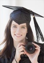 Portrait of young woman in graduation gown holding camera. Photographe : Jamie Grill