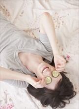 Young woman lying in bed with cucumber face mask. Photographe : Jamie Grill