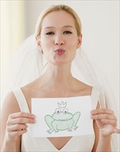 Portrait of young bride blowing kiss and holding drawing of frog prince. Photographe : Jamie Grill