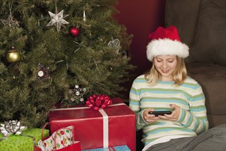 Teenage girl (13-15) sitting in front of Christmas tree and text messaging. Photographe : Sarah M.