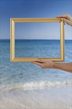 Hands holding a picture frame at a beach.
