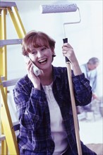 Woman talking on phone while holding a paint roller. Photographe : Rob Lewine