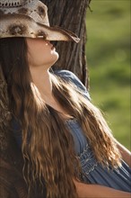 Cowgirl leaning against tree with her hat covering her face. Photo : Mike Kemp