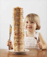 Young girl sitting behind a tall stack of pancakes. Photo. FBP