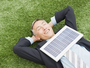 Happy businessman lying on grass with eyes closed and hands behind head and with solar panel on