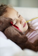 Sad girl (10-11) lying in bed with teddybear and thermometer in mouth. Photo : Momentimages