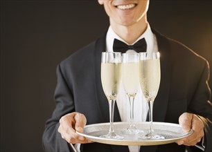 Waiter wearing bow tie and holding tray with champagne flutes, studio shot. Photo : Jamie Grill